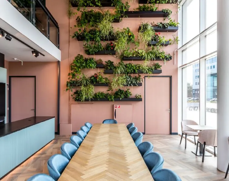 Vergaderruimte aan het John M. Keynesplein met een lange houten tafel, blauwe stoelen en een groene plantenwand tegen een roze muur.